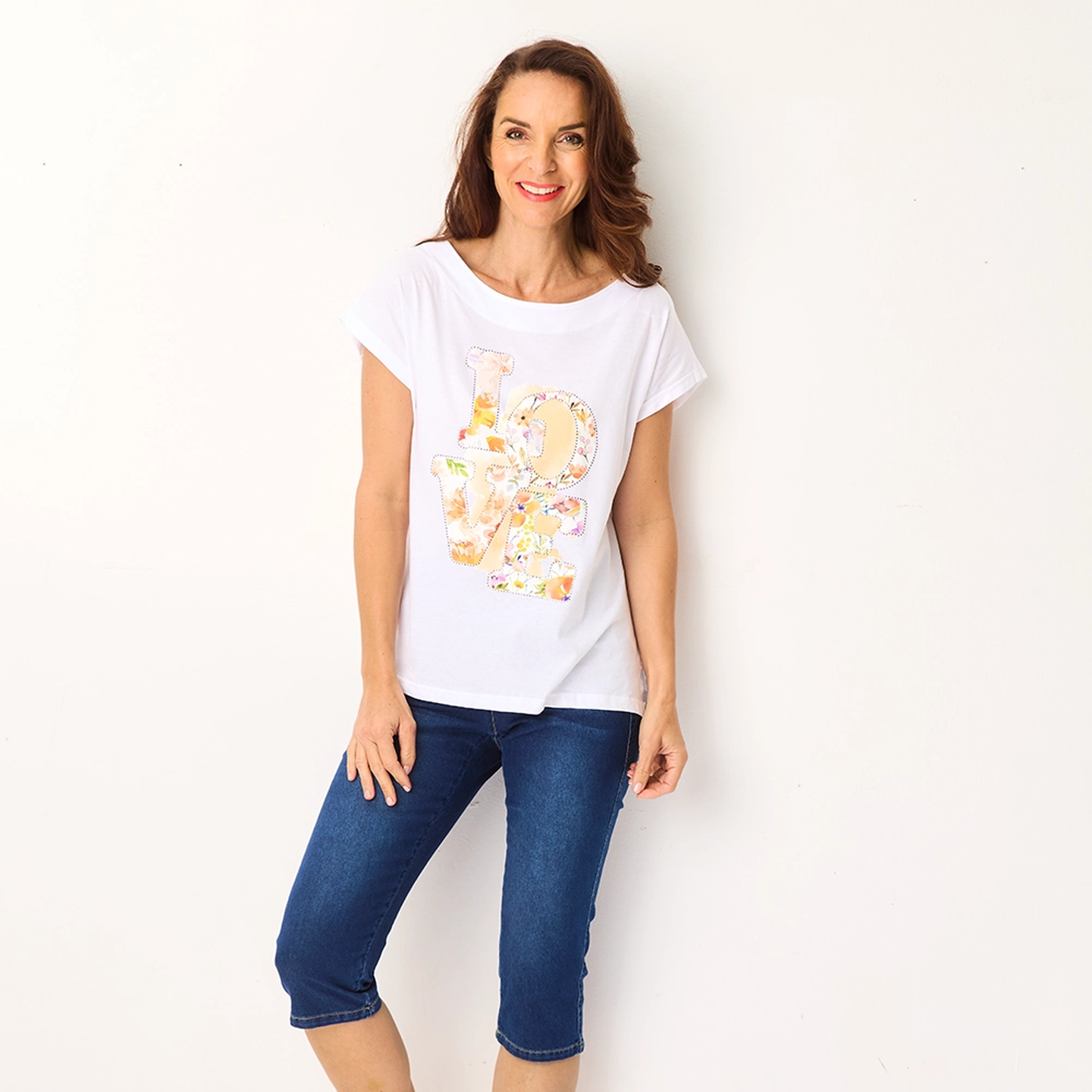 Frau lächelt, trägt ein weißes T-Shirt mit dem Schriftzug „LOVE” in bunten Buchstaben und eine blaue Caprihose, steht vor einem schlichten weißen Hintergrund.
