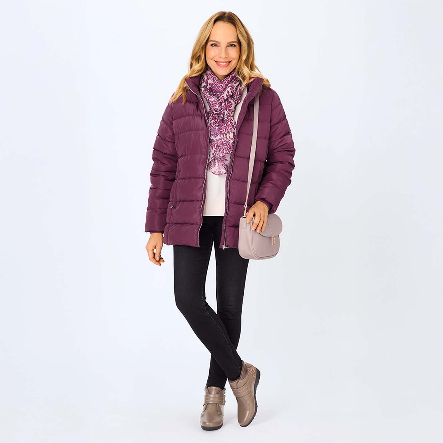 Eine Frau in einer lila Daunenjacke, einem geblümten Schal, einer schwarzen Hose und taupefarbenen Schuhen, die eine helllila Handtasche in der Hand hält und vor einem schlichten Hintergrund lächelt.