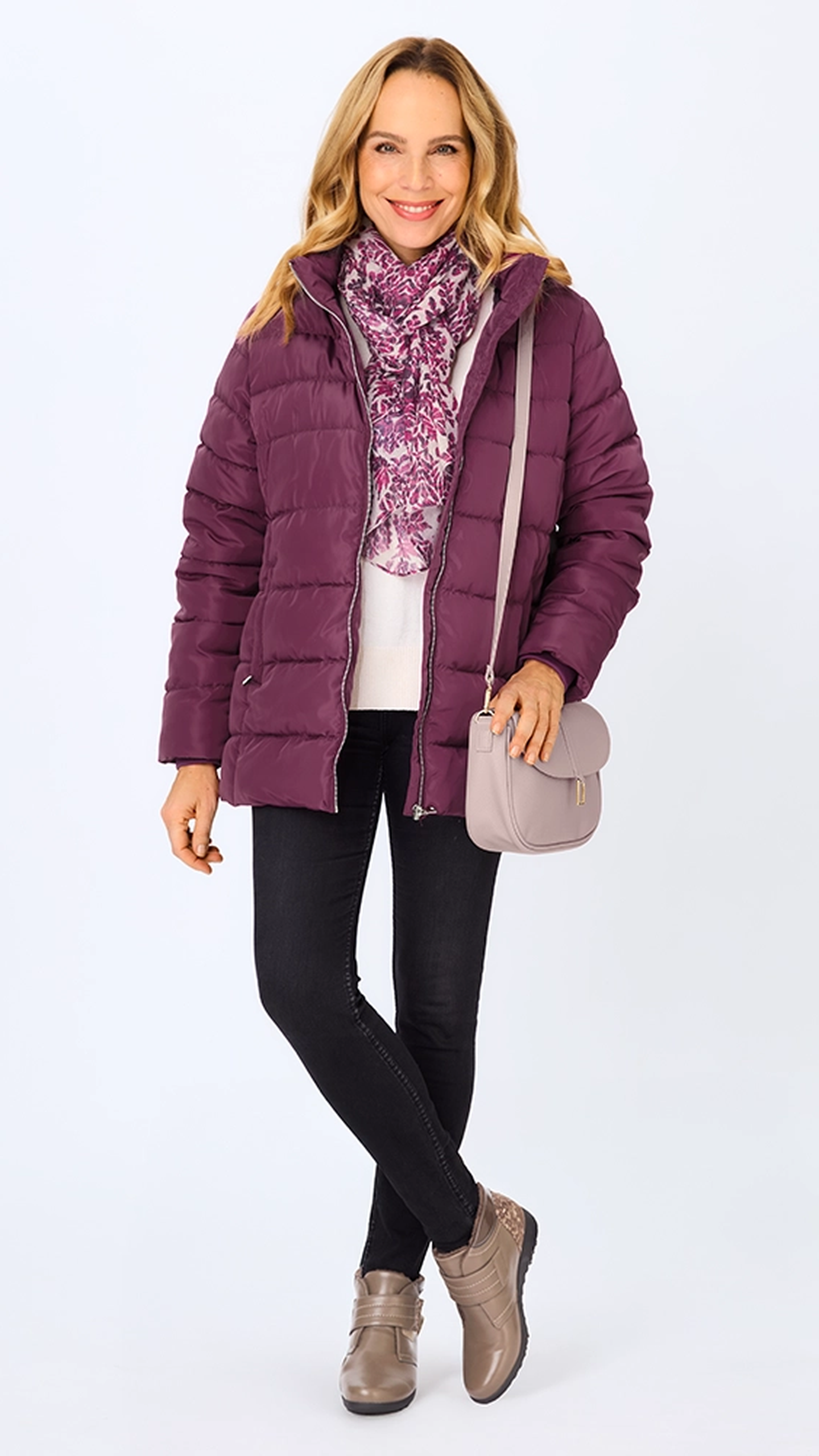 Eine Frau in einer lila Daunenjacke, einem geblümten Schal, einer schwarzen Hose und taupefarbenen Schuhen, die eine helllila Handtasche in der Hand hält und vor einem schlichten Hintergrund lächelt.