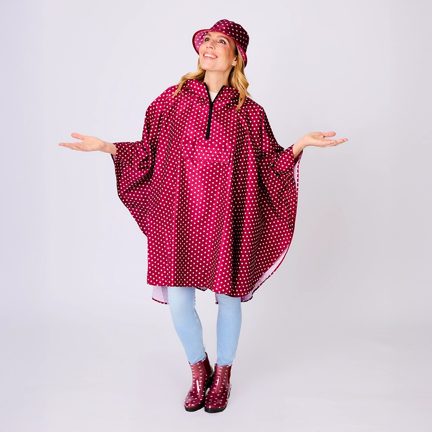 Eine lächelnde Frau, die einen gepunkteten Regenponcho und einen dazu passenden Hut trägt, dazu hellblaue Jeans und weinrote Stiefel, die Arme in einer verspielten Pose ausgestreckt.