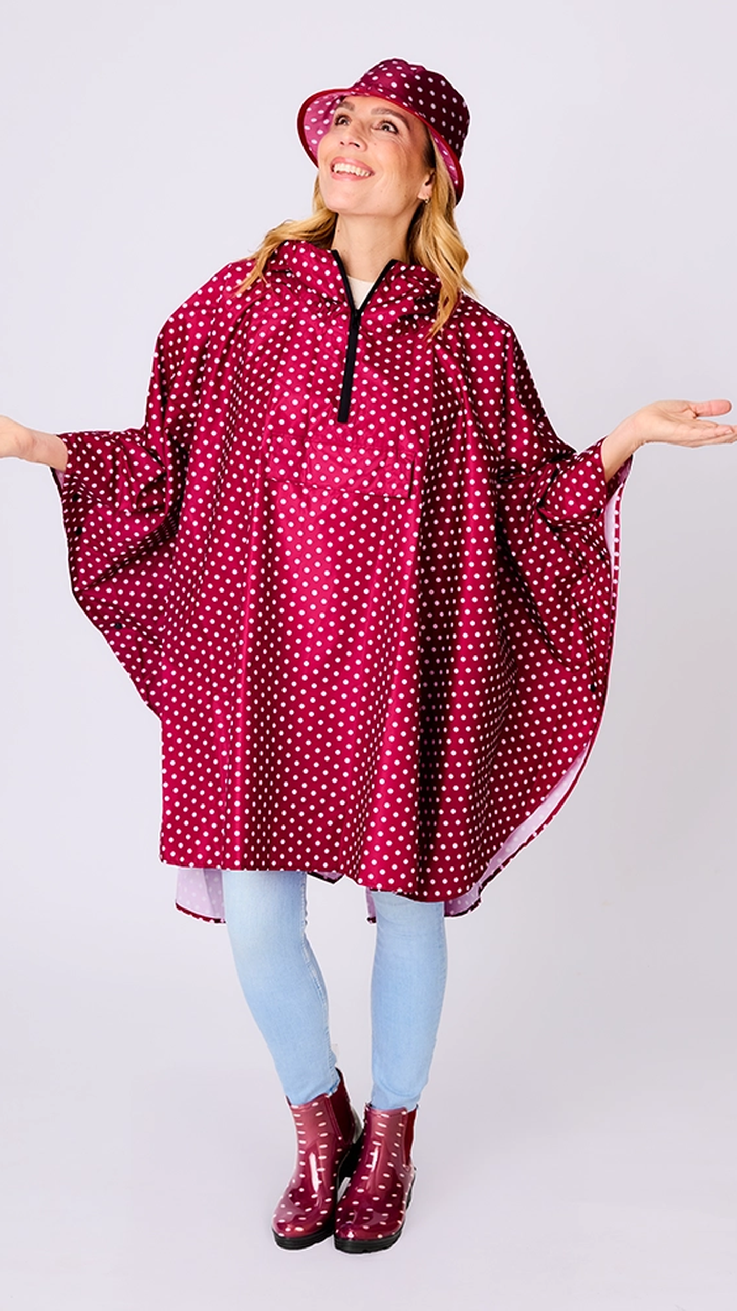 Eine lächelnde Frau, die einen gepunkteten Regenponcho und einen dazu passenden Hut trägt, dazu hellblaue Jeans und weinrote Stiefel, die Arme in einer verspielten Pose ausgestreckt.
