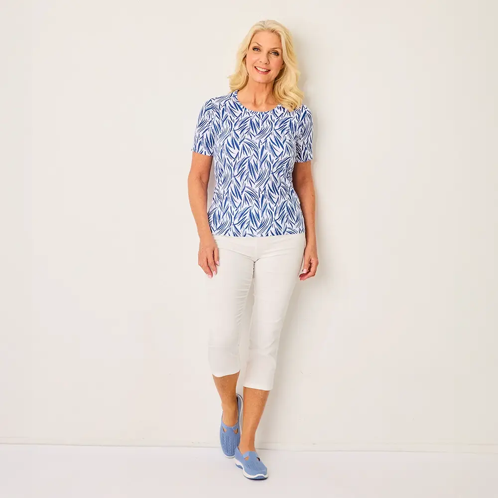 Frau in einem blau gemusterten Oberteil und weißer Caprihose steht lächelnd vor einem schlichten Hintergrund und trägt blaue Slipper.