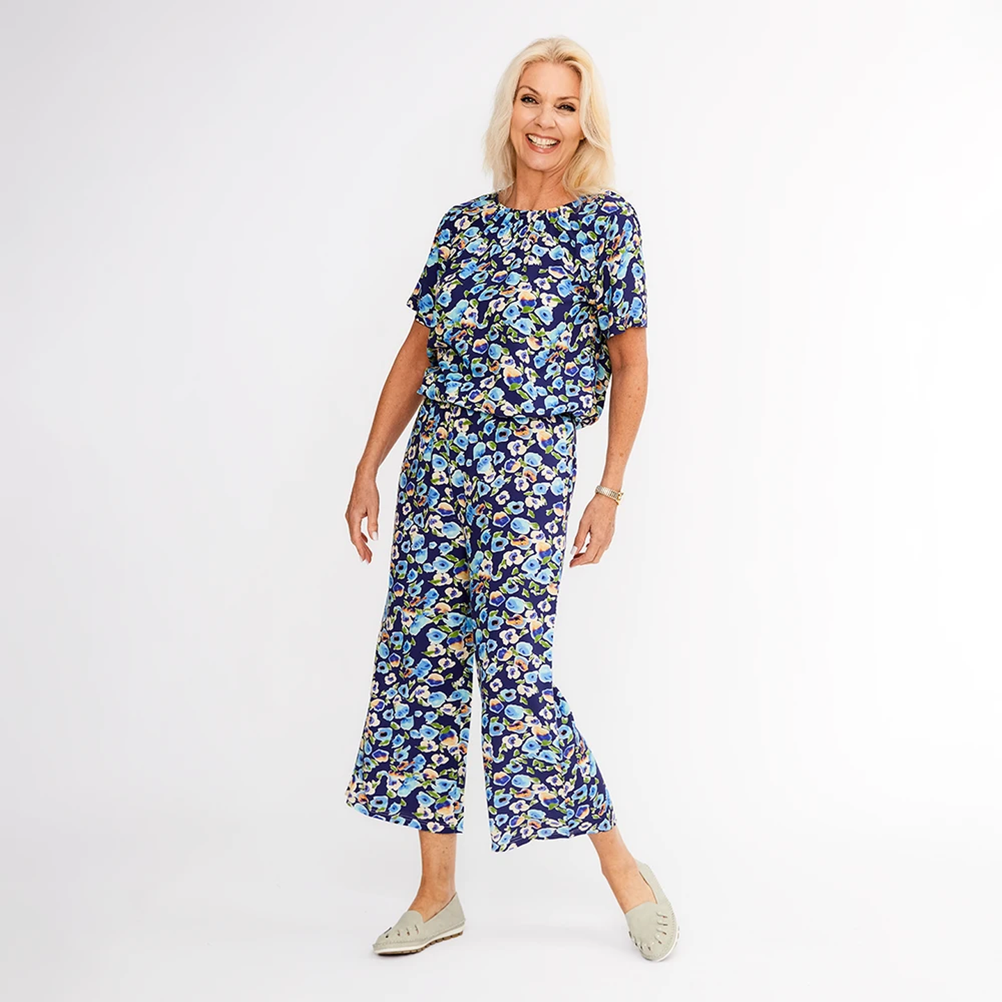 Eine lächelnde Frau in einem blauen, geblümten Outfit mit weit geschnittenen Hosen posiert vor einem schlichten weißen Hintergrund.