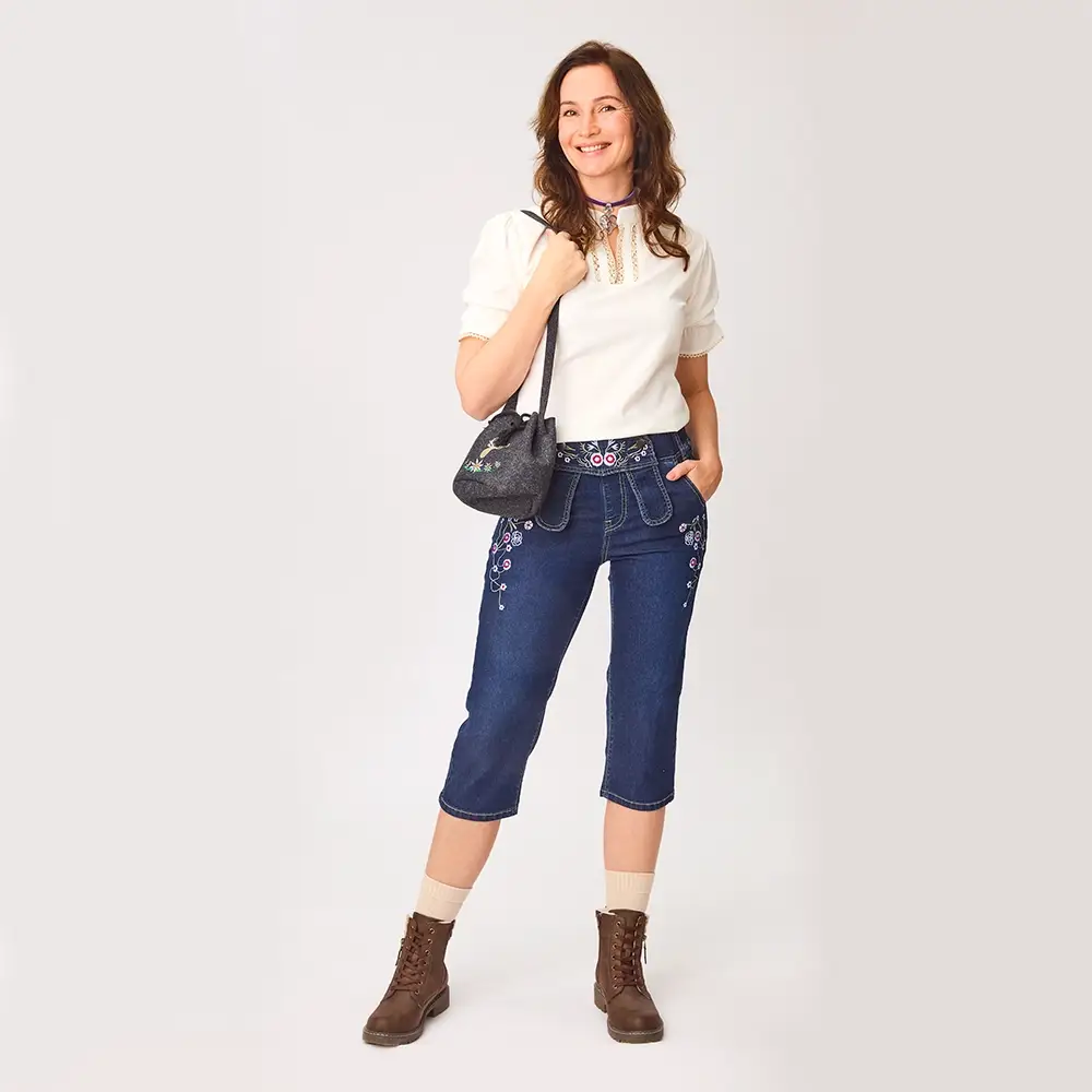 Eine Frau in einer weißen Bluse und einer bestickten blauen Caprihose, die eine schwarze Handtasche hält, lächelt vor einem einfarbigen Hintergrund.