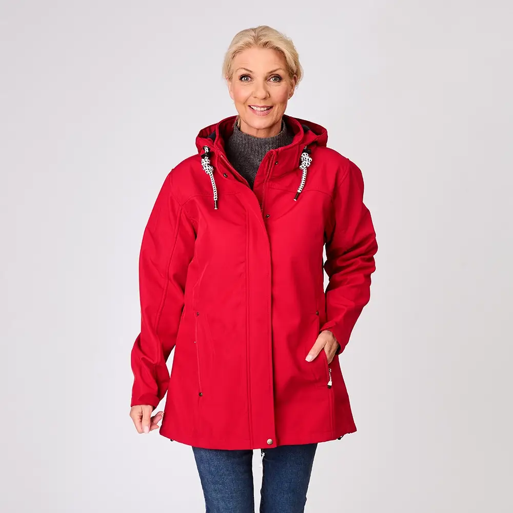 Frau in einer roten Kapuzenjacke und blauen Jeans lächelt vor einem einfarbigen Hintergrund.