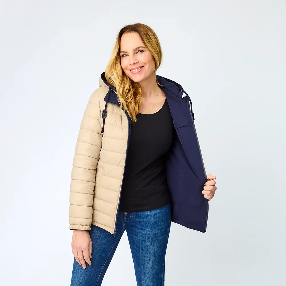 Lächelnde Frau, die eine beige-marineblaue Wendejacke und Jeans trägt und die Jacke öffnet, um das Innenfutter zu zeigen.