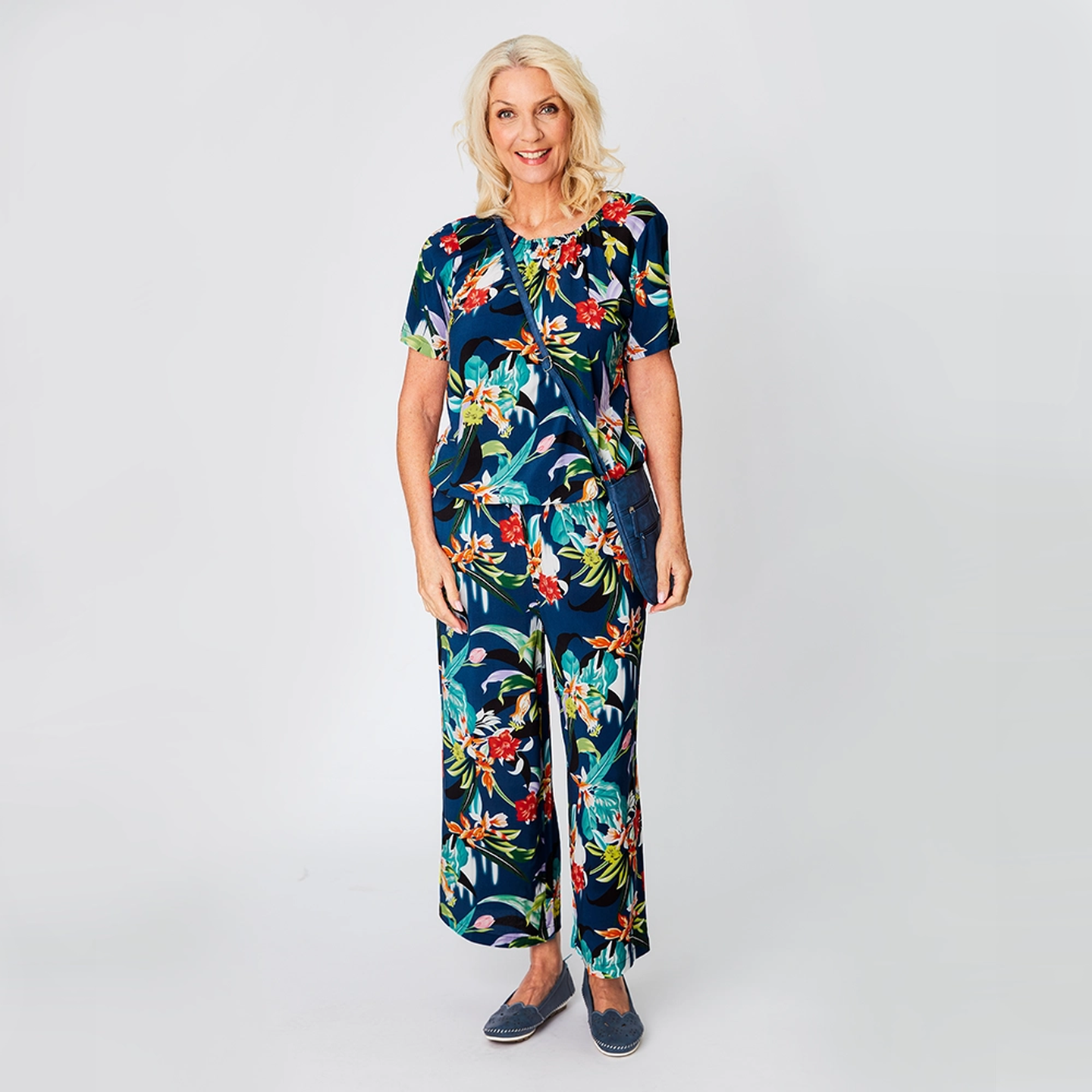 Eine Frau in einem farbenfrohen Blumenoutfit steht vor einem hellen Hintergrund und lächelt. Sie trägt eine dunkle Umhängetasche und blaue Schuhe.