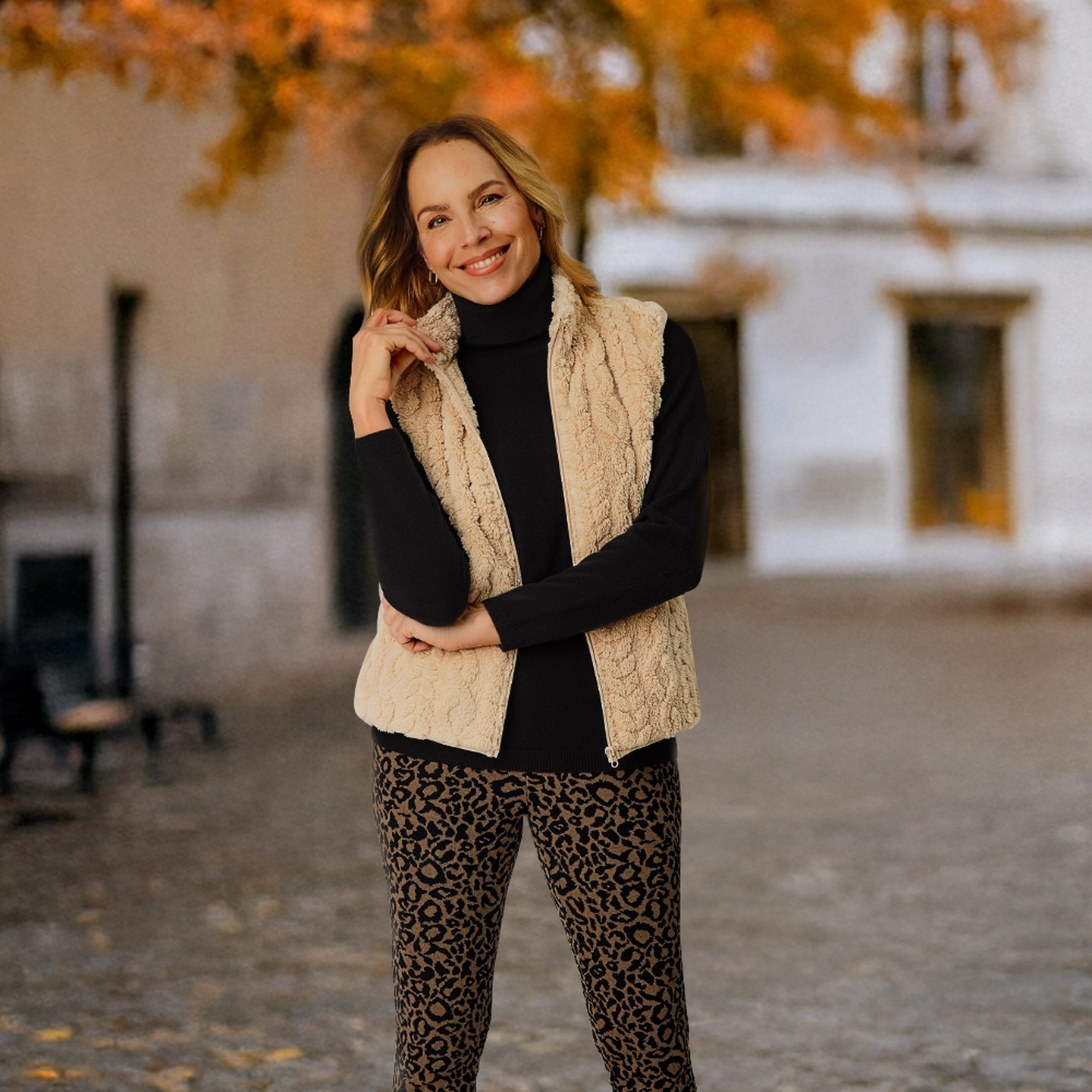 Eine Frau lächelt. Sie trägt eine beige Weste, einen schwarzen Rollkragenpullover und eine Hose mit Leopardenmuster und steht auf einer Straße, im Hintergrund ist Herbstlaub zu sehen.
