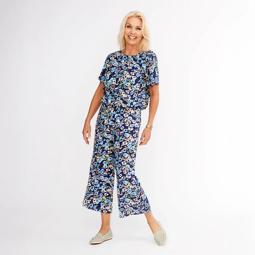Frau in einem blauen Blumenoutfit mit weiten Hosen und kurzärmeligem Oberteil, lächelnd und vor einem weißen Hintergrund stehend.
