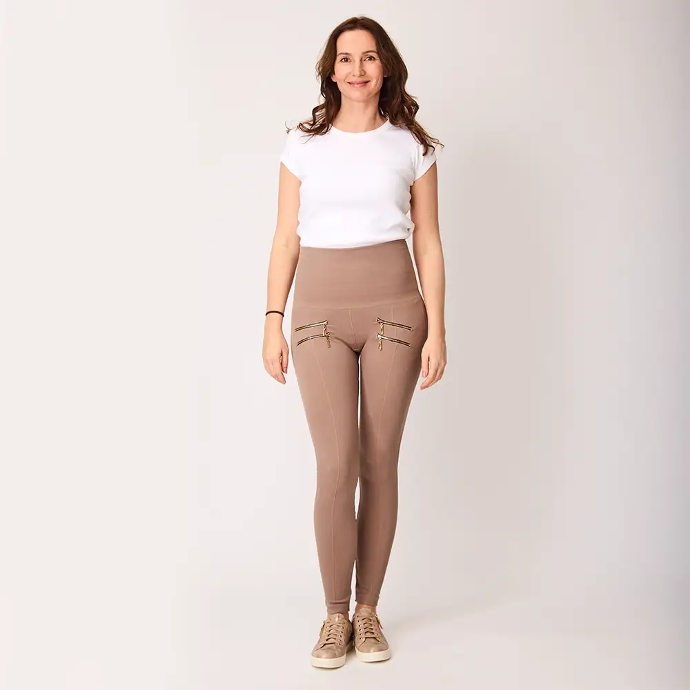 Frau in einem weißen T-Shirt und beigen Leggings mit Reißverschlussdetails, die vor einem einfarbigen Hintergrund steht und lächelt.