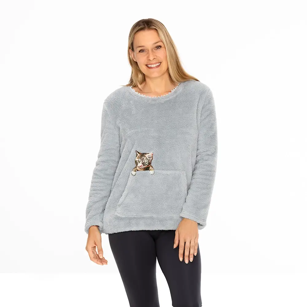 Lächelnde Frau, die einen hellgrauen, flauschigen Pullover mit einem Katzenmotiv auf der Vordertasche trägt, dazu schwarze Leggings, vor einem weißen Hintergrund.