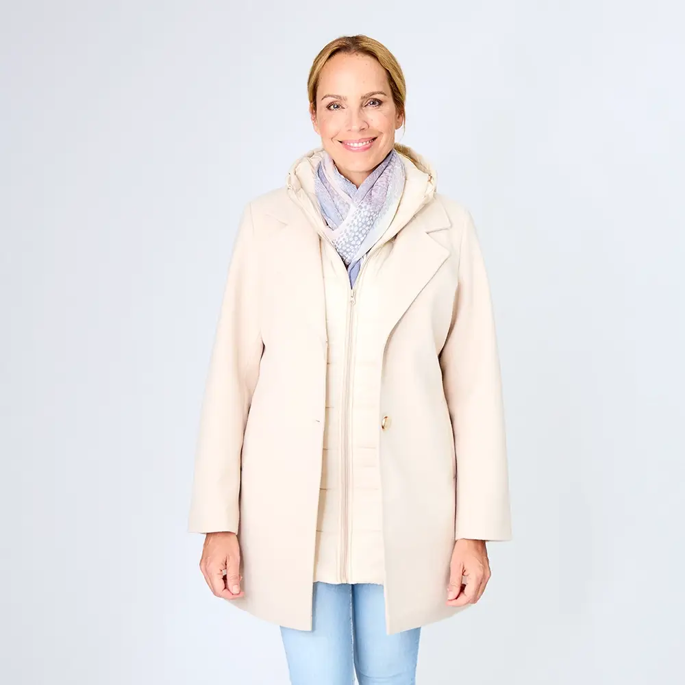 Frau lächelt, trägt einen hellbeigen Mantel über einer Daunenjacke, dazu einen grauen Schal und hellblaue Jeans, vor einem schlichten weißen Hintergrund.
