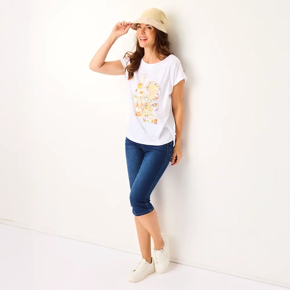 Frau lächelt, trägt einen beigen Hut, ein weißes bedrucktes T-Shirt, blaue Capri-Jeans und weiße Sneakers, und lehnt an einer weißen Wand.