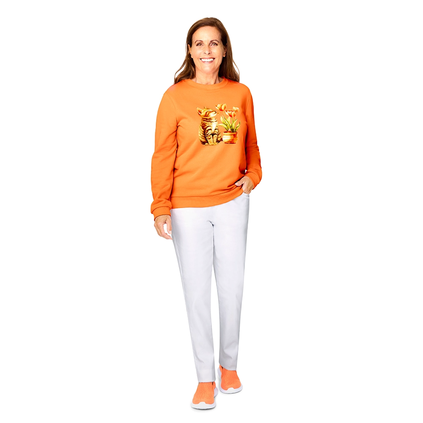 Eine Frau in einem orangefarbenen Pullover mit Katzenmotiv, weißen Hosen und orangefarbenen Schuhen steht lächelnd vor einem weißen Hintergrund.