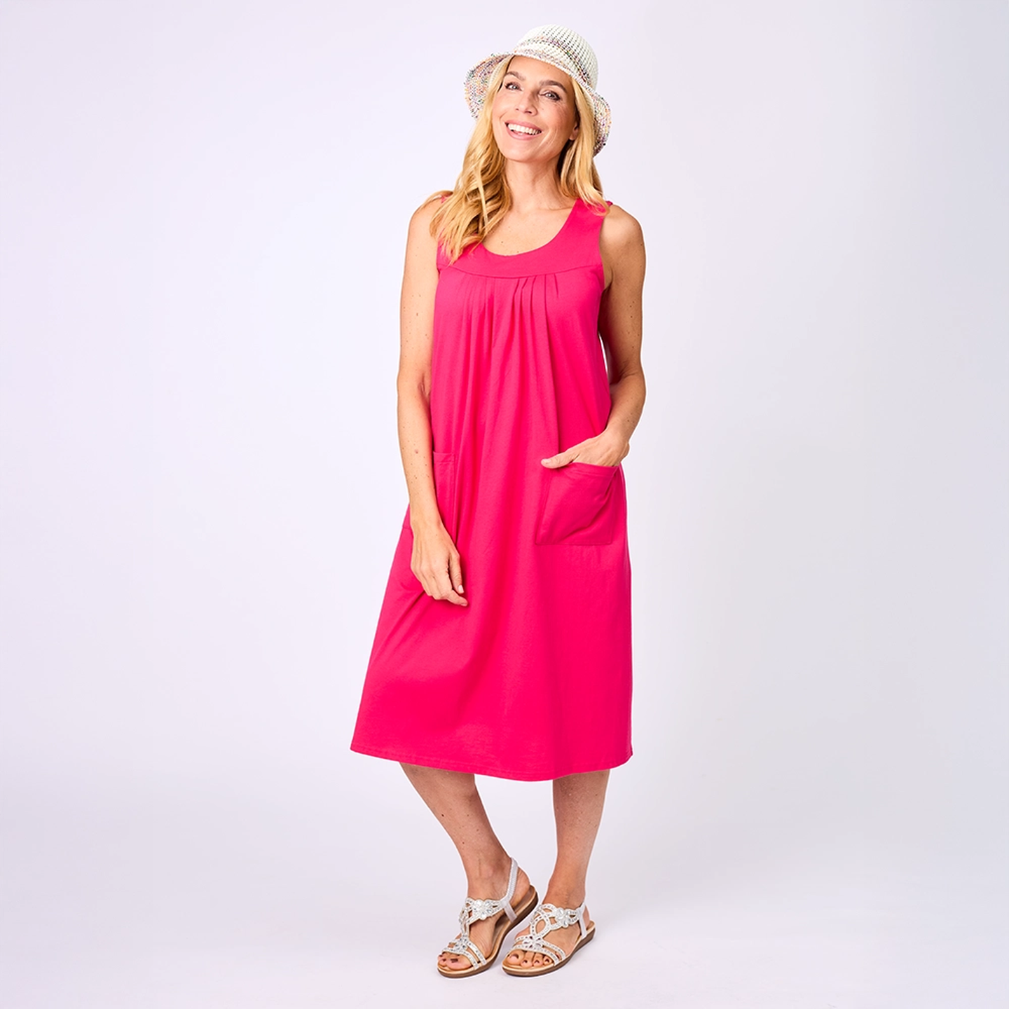 Eine Frau in einem rosa, ärmellosen Kleid und einem Sonnenhut, lächelnd, mit einer Hand in der Tasche, vor einem schlichten Hintergrund stehend.