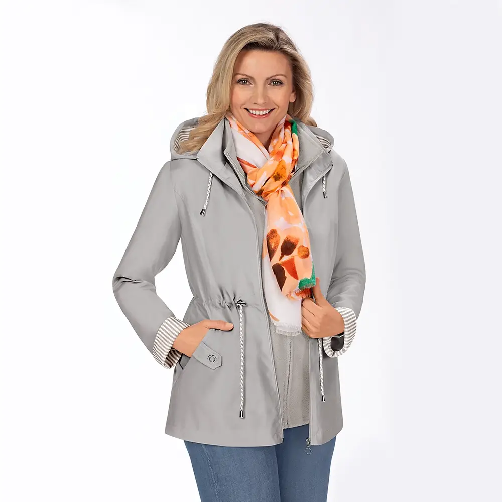 Lächelnde Frau in einer grauen Kapuzenjacke und Jeans, mit einem orange-weißen Schal, vor einem schlichten weißen Hintergrund stehend.