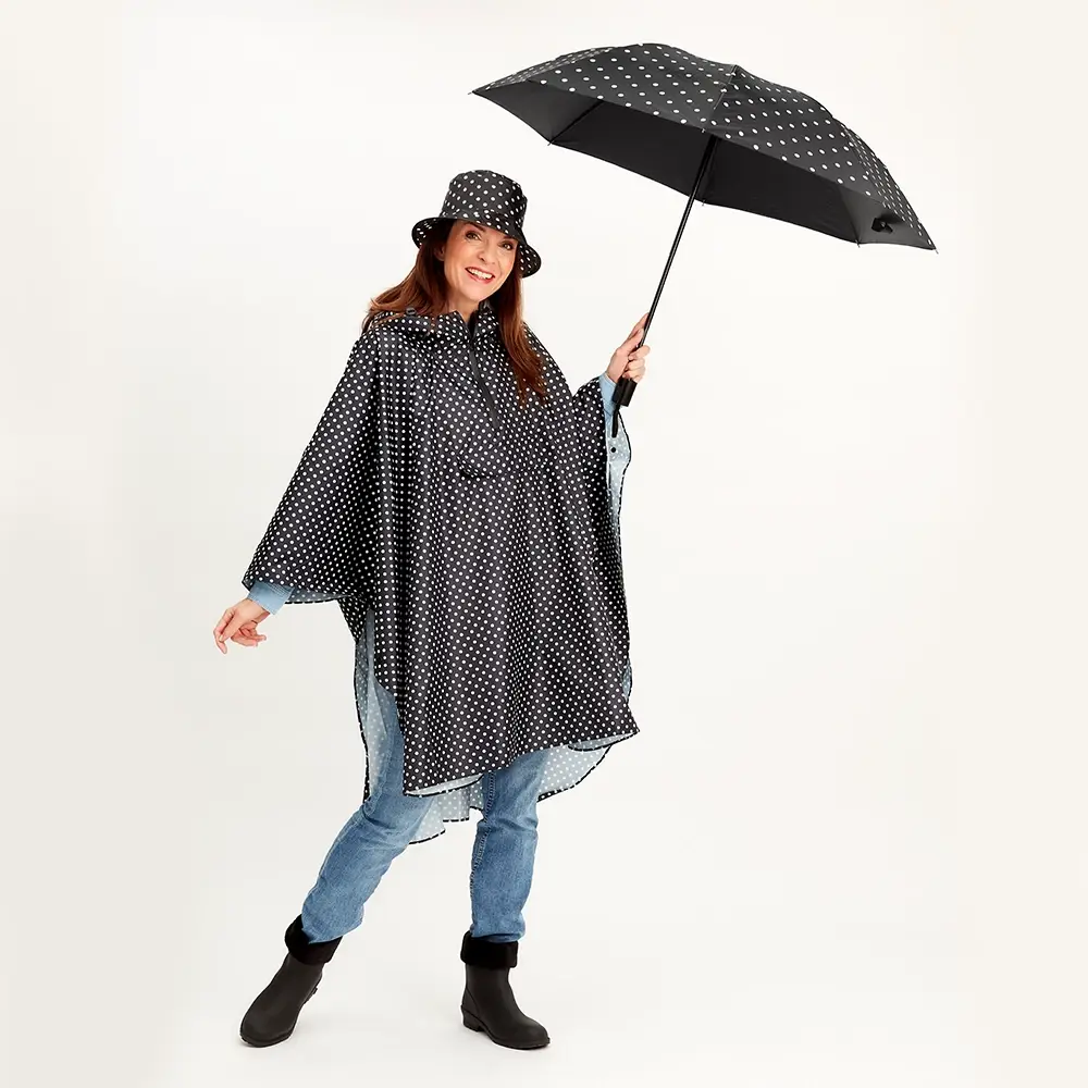 Frau in einem gepunkteten Regenmantel und Hut, die einen passenden Regenschirm hält, Jeans und schwarze Stiefel trägt und vor einem einfarbigen Hintergrund lächelt.