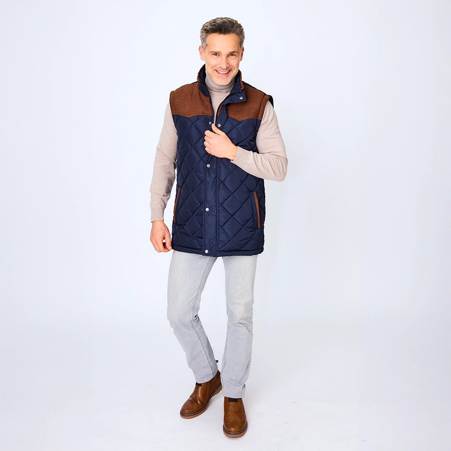 Ein Mann in einer gesteppten, marineblauen Weste mit braunen Akzenten, einem beigen Rollkragenpullover, hellgrauen Hosen und braunen Stiefeln, der vor einem weißen Hintergrund lächelt.