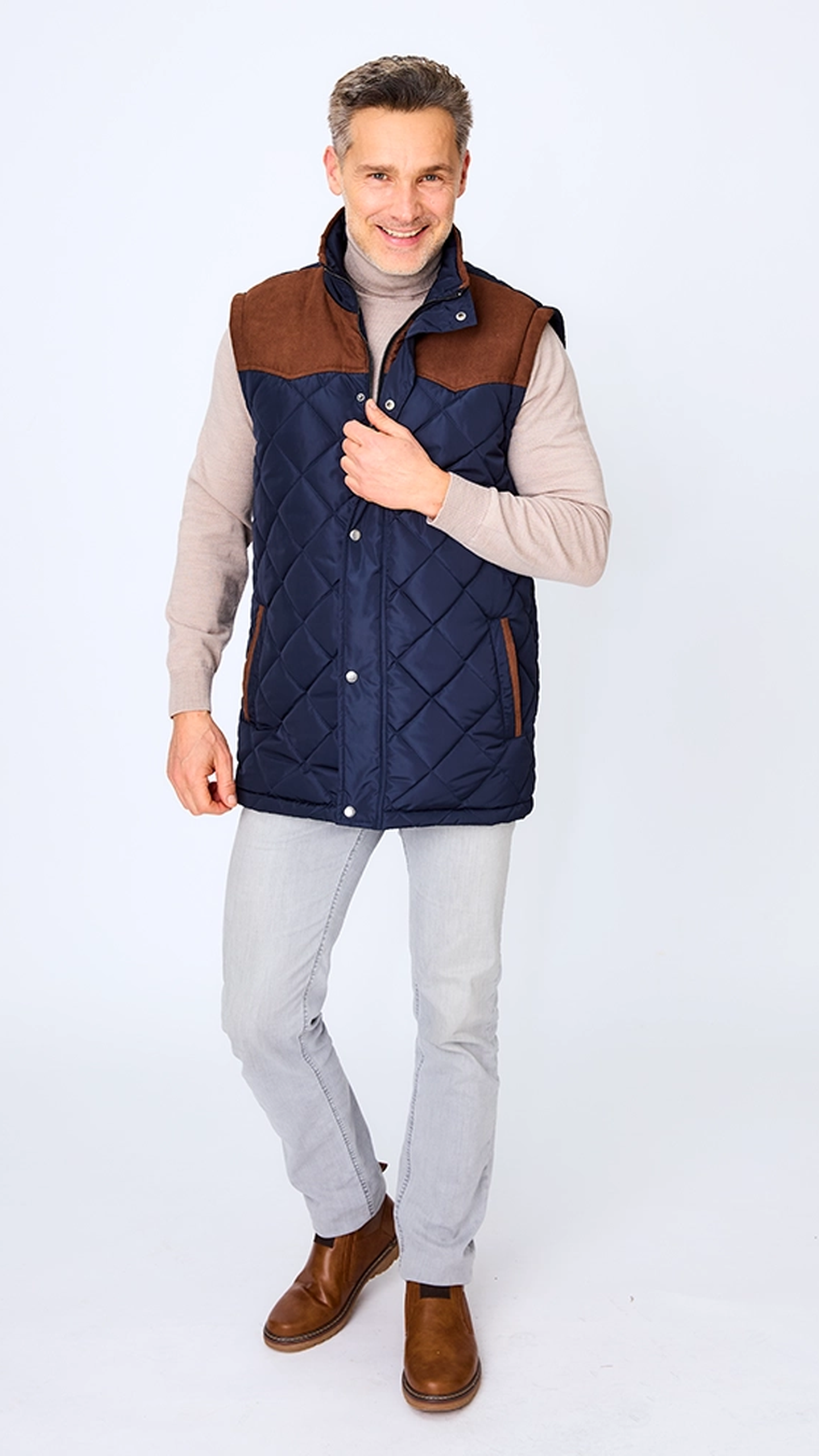 Ein Mann in einer gesteppten, marineblauen Weste mit braunen Akzenten, einem beigen Rollkragenpullover, hellgrauen Hosen und braunen Stiefeln, der vor einem weißen Hintergrund lächelt.
