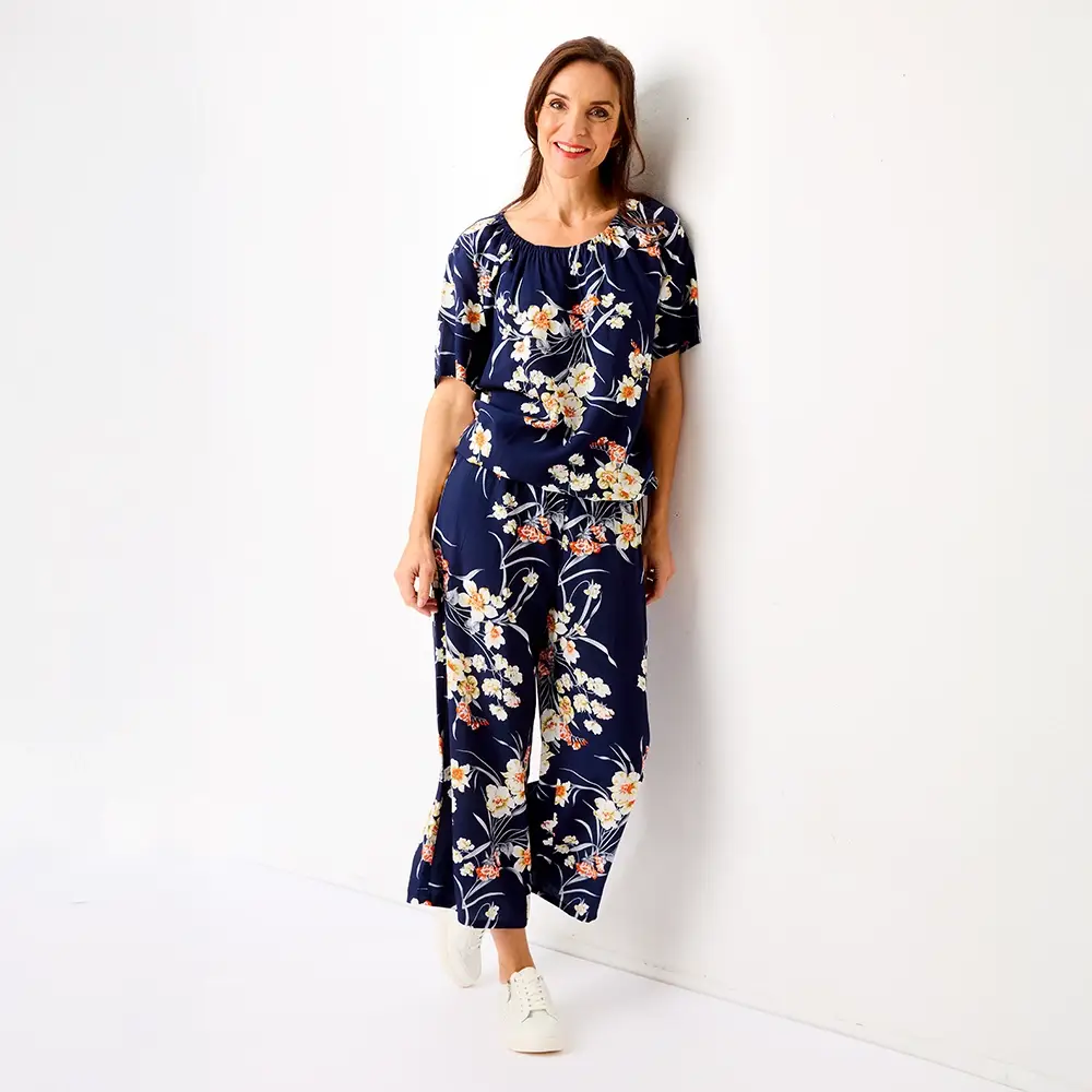 Frau in einem blumigen, navyfarbenen Jumpsuit und weißen Sneakers lehnt an einer weißen Wand, lächelt und hat die Hände in den Taschen.