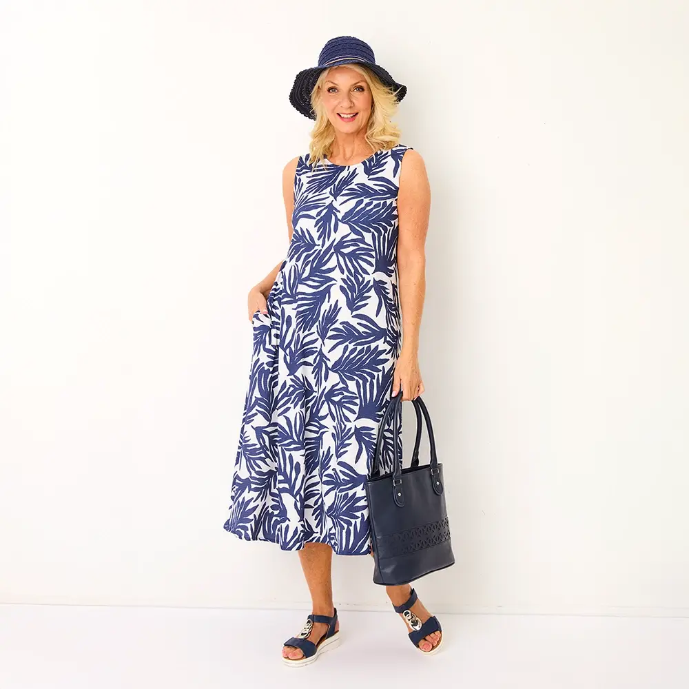 Frau in einem blau-weiß gemusterten Kleid mit Blattmotiv, marineblauem Hut und Sandalen, hält eine passende Handtasche und steht vor einem weißen Hintergrund.