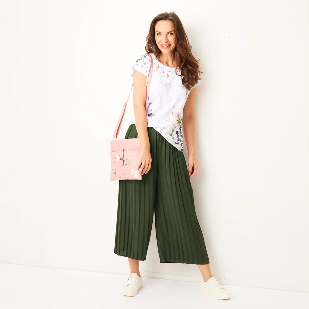 Frau in einem blumigen Oberteil, grünen, plissierten Hosen und weißen Sneakers, hält eine rosa Handtasche und lächelt vor einem weißen Hintergrund.