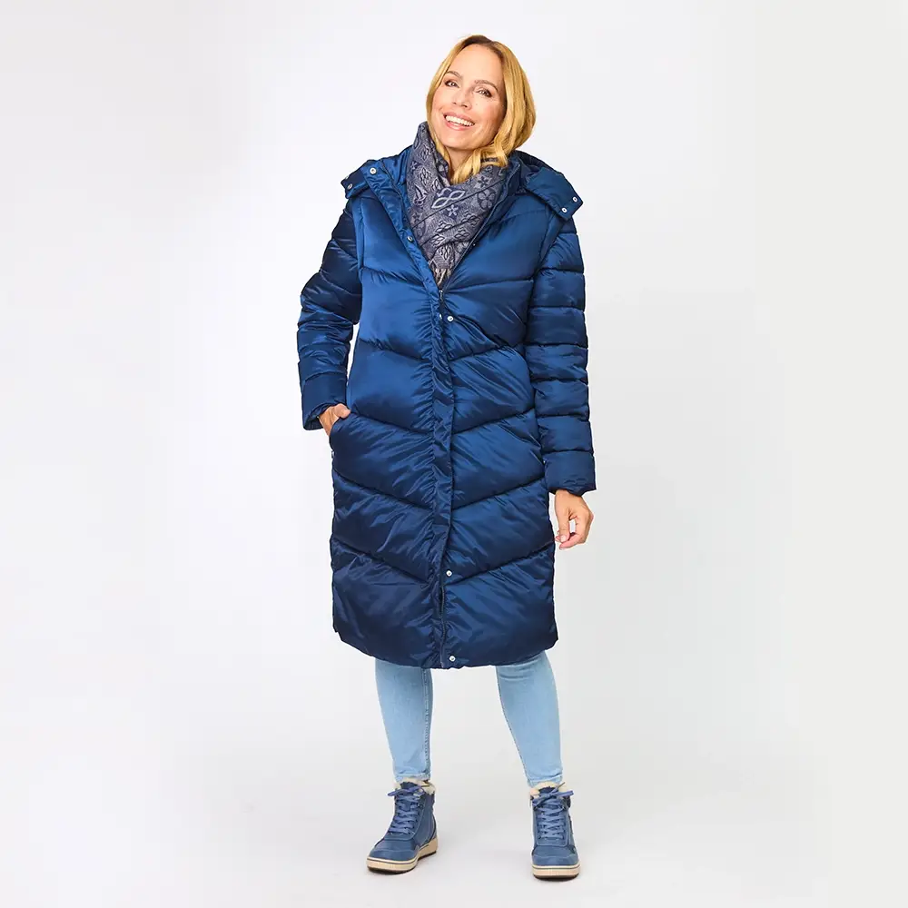 Frau in einem blauen Daunenmantel und Jeans, lächelnd, vor einem weißen Hintergrund stehend. Sie trägt blaue Schuhe und einen gemusterten Schal.