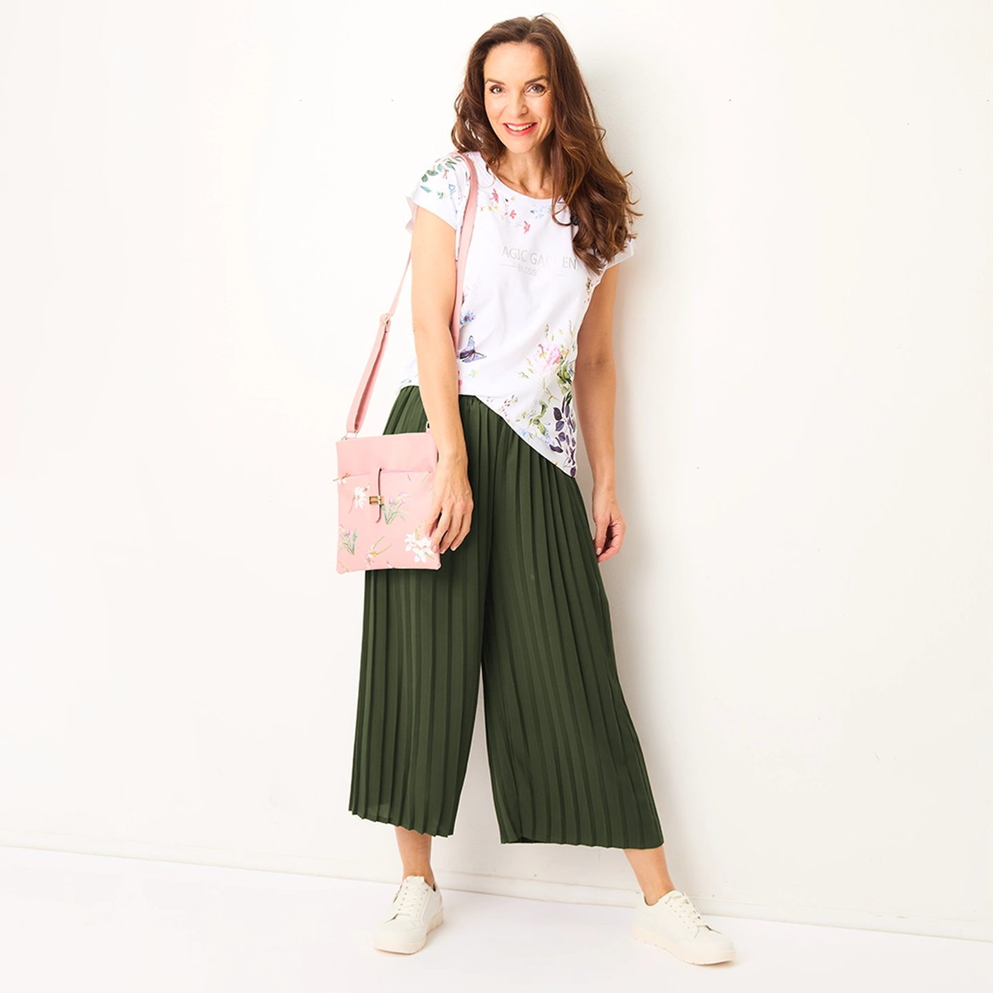 Frau in einem geblümten Oberteil, grüner Plisseehose und weißen Sneakers, die eine rosa geblümte Tasche vor einem weißen Hintergrund hält.