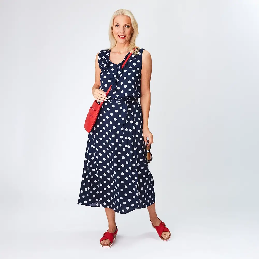 Frau in einem marineblauen gepunkteten Kleid mit roter Tasche und Schuhen, mit Sonnenbrille, steht vor einem schlichten weißen Hintergrund.