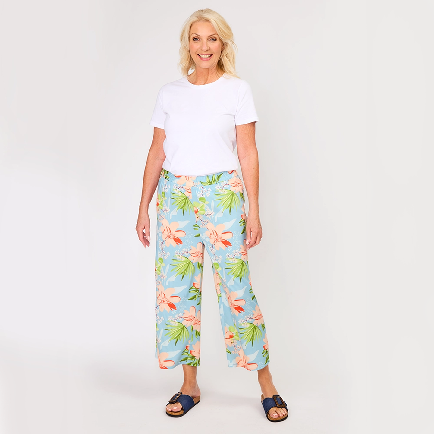Eine lächelnde Frau in einem weißen T-Shirt und einer Hose mit Blumenmuster steht vor einem einfarbigen Hintergrund und trägt blaue Sandalen.