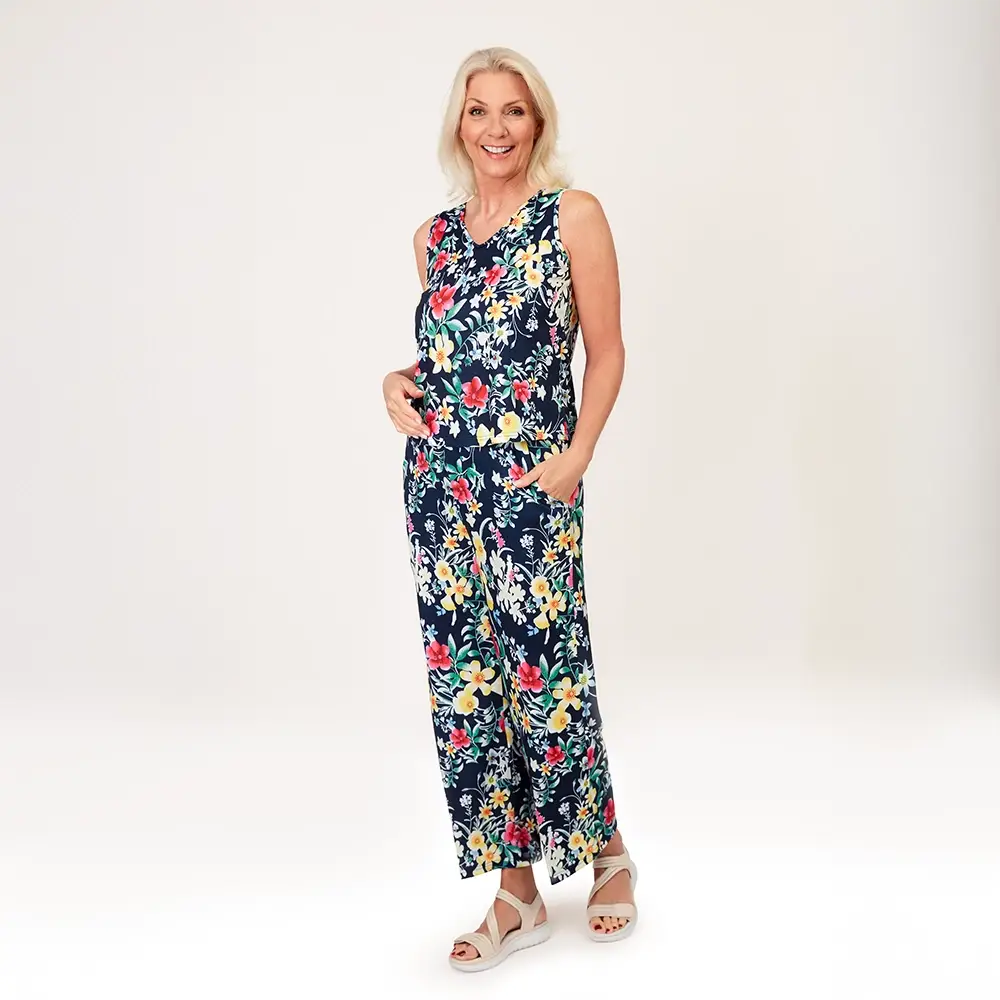 Lächelnde Frau in einem ärmellosen, blumigen Jumpsuit mit den Händen in den Taschen, trägt weiße Sandalen und steht vor einem schlichten Hintergrund.