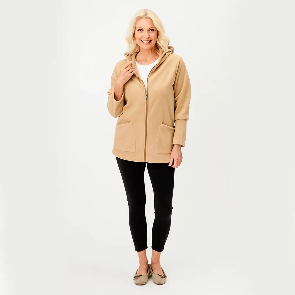 Frau in einem beigen Kapuzenpullover mit Reißverschluss und schwarzen Leggings, lächelnd, vor einem schlichten weißen Hintergrund stehend.