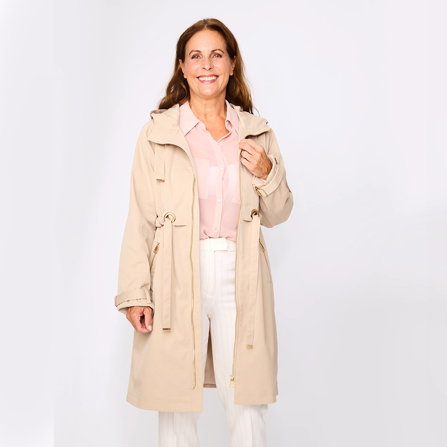 Lächelnde Frau in einem beigen Mantel über einer rosa Bluse und einer weißen Hose, vor einem schlichten weißen Hintergrund stehend.