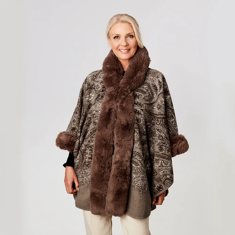 Frau in einem gemusterten Poncho mit Pelzbesatz, lächelnd vor einem einfarbigen Hintergrund.