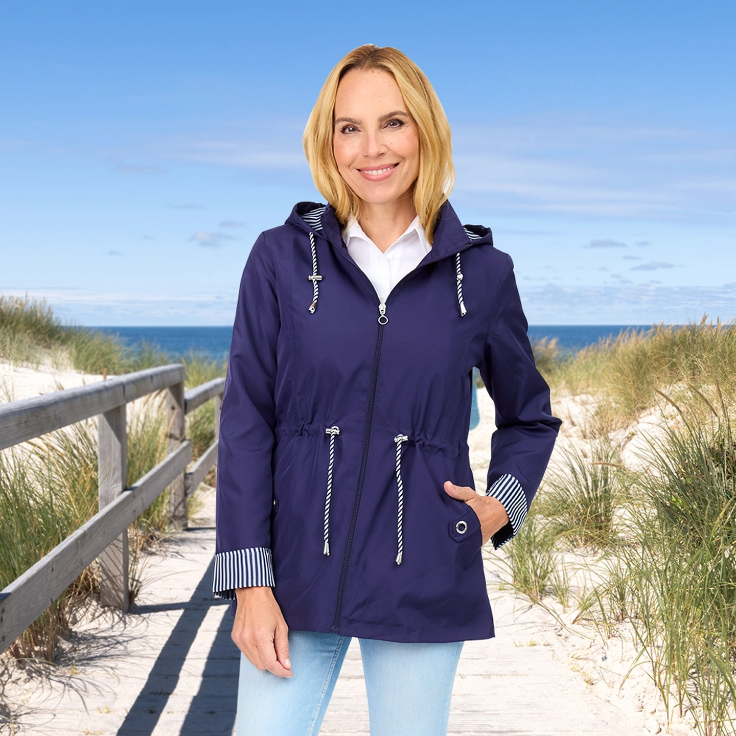 Eine Frau in einer violetten Jacke steht auf einem sandigen Strandweg, im Hintergrund sind grasbewachsene Dünen und das Meer zu sehen, über ihr erstreckt sich ein strahlend blauer Himmel.