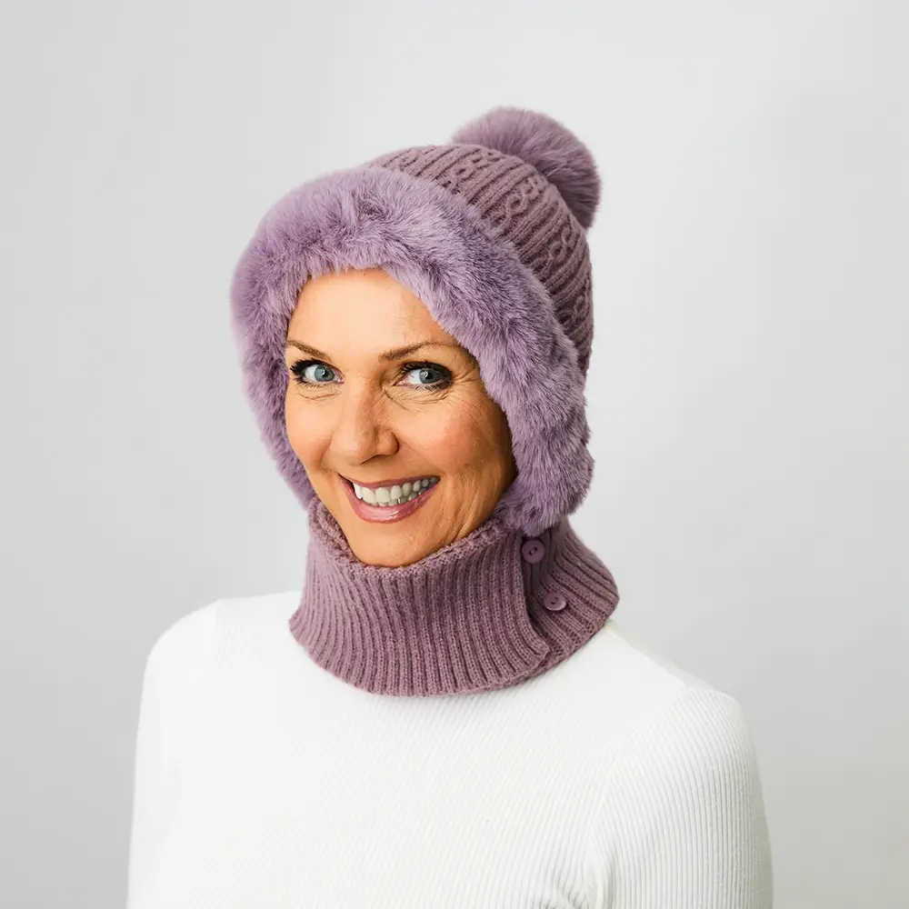 Lächelnde Person mit einer violetten Strickmütze mit Kunstfellbesatz und passendem Halswärmer vor einem schlichten weißen Hintergrund.