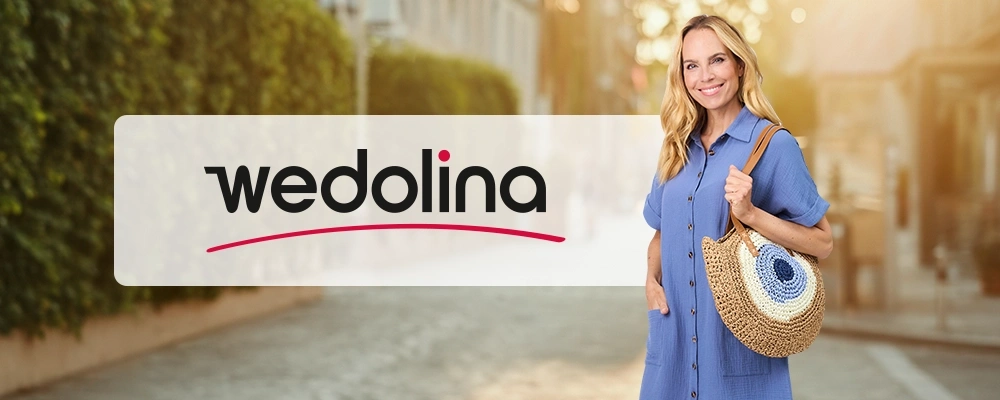 Eine Frau in einem blauen Kleid, die eine gewebte Tasche in der Hand hält, steht lächelnd auf einer sonnigen Straße neben dem Wedolina-Logo.