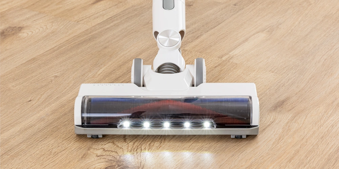 Ein kabelloser Staubsaugerkopf mit LED-Leuchten wird auf einen Holzboden gestellt. Die Leuchten beleuchten den Bereich vor dem Staubsauger.