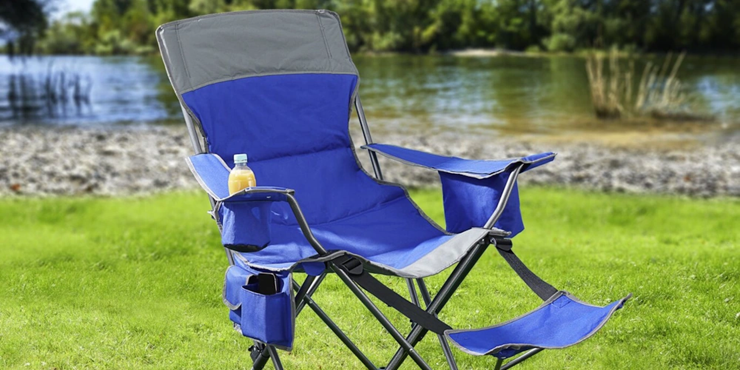 Ein blauer Klapp-Campingstuhl mit Kopfstütze und Getränkehalter steht auf dem Rasen am Seeufer; er verfügt über eine Fußstütze und eine Aufbewahrungstasche.