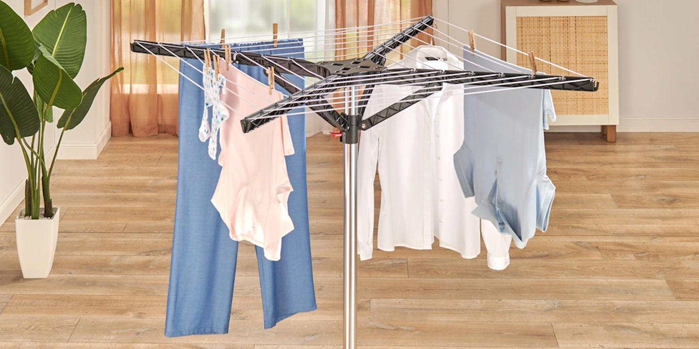 Eine Wäschespinne, an der verschiedene Kleidungsstücke, darunter Hemden und Hosen, auf einem Holzboden in der Nähe einer Pflanze zum Trocknen aufgehängt sind.