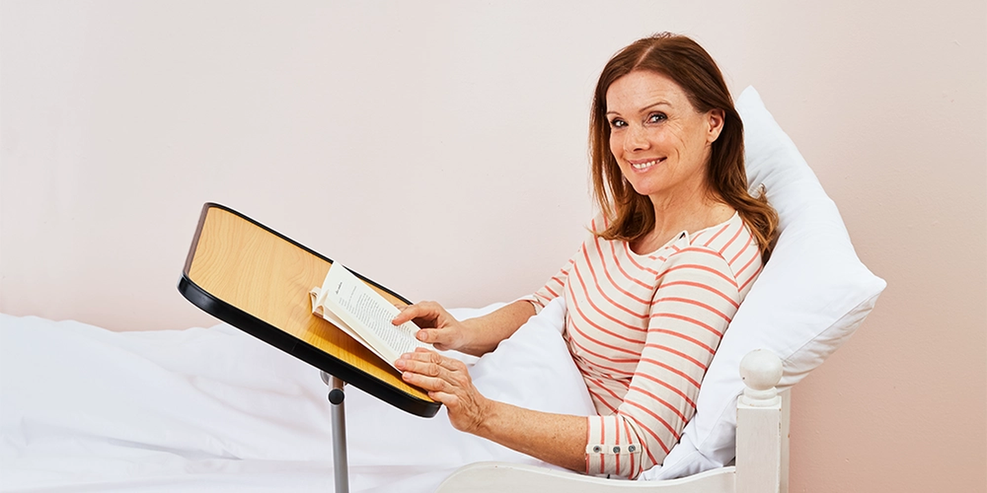 Eine Frau in einem gestreiften Shirt lächelt, während sie im Bett auf dem Beistelltisch ein Buch liest; sie lehnt an weißen Kissen und einer hellrosa Wand.