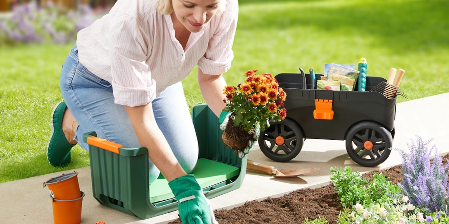 Eine Person kniet auf einem Gartensitz und pflanzt Blumen. Daneben steht ein Gartenwagen mit Werkzeugen und Gartenbedarf. Ein sonniger Tag auf einer Rasenfläche.