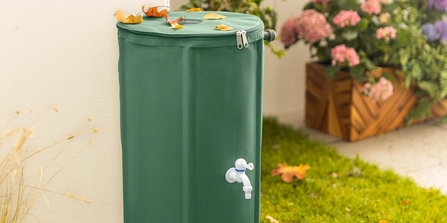 Ein grüner Regensammelbehälter mit einem Zapfhahn, umgeben von Laub, in der Nähe eines Gartens mit rosa und violetten Blumen in einem hölzernen Pflanzkübel.