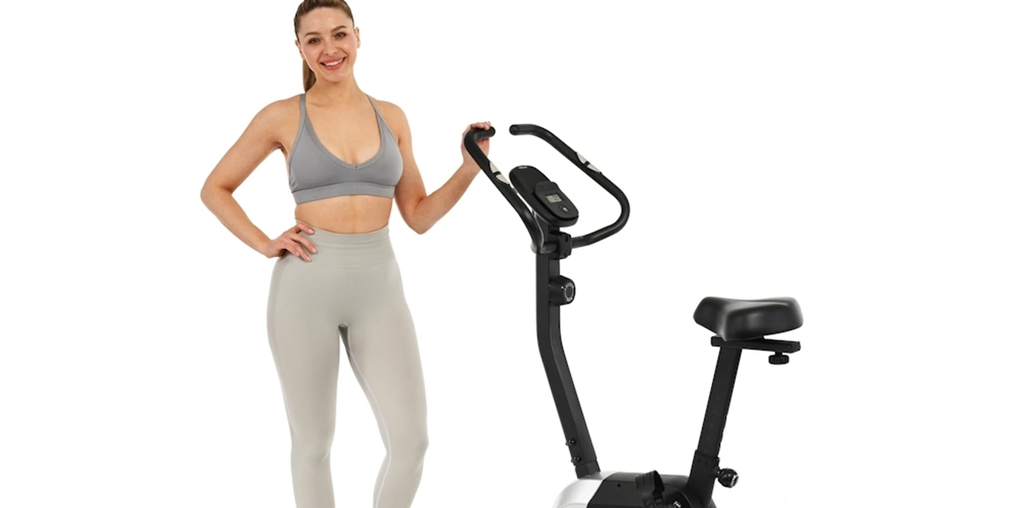 Eine Frau in Sportkleidung steht neben einem stationären Heimtrainer, lächelt und stützt die Hand auf den Lenker, vor einem weißen Hintergrund.