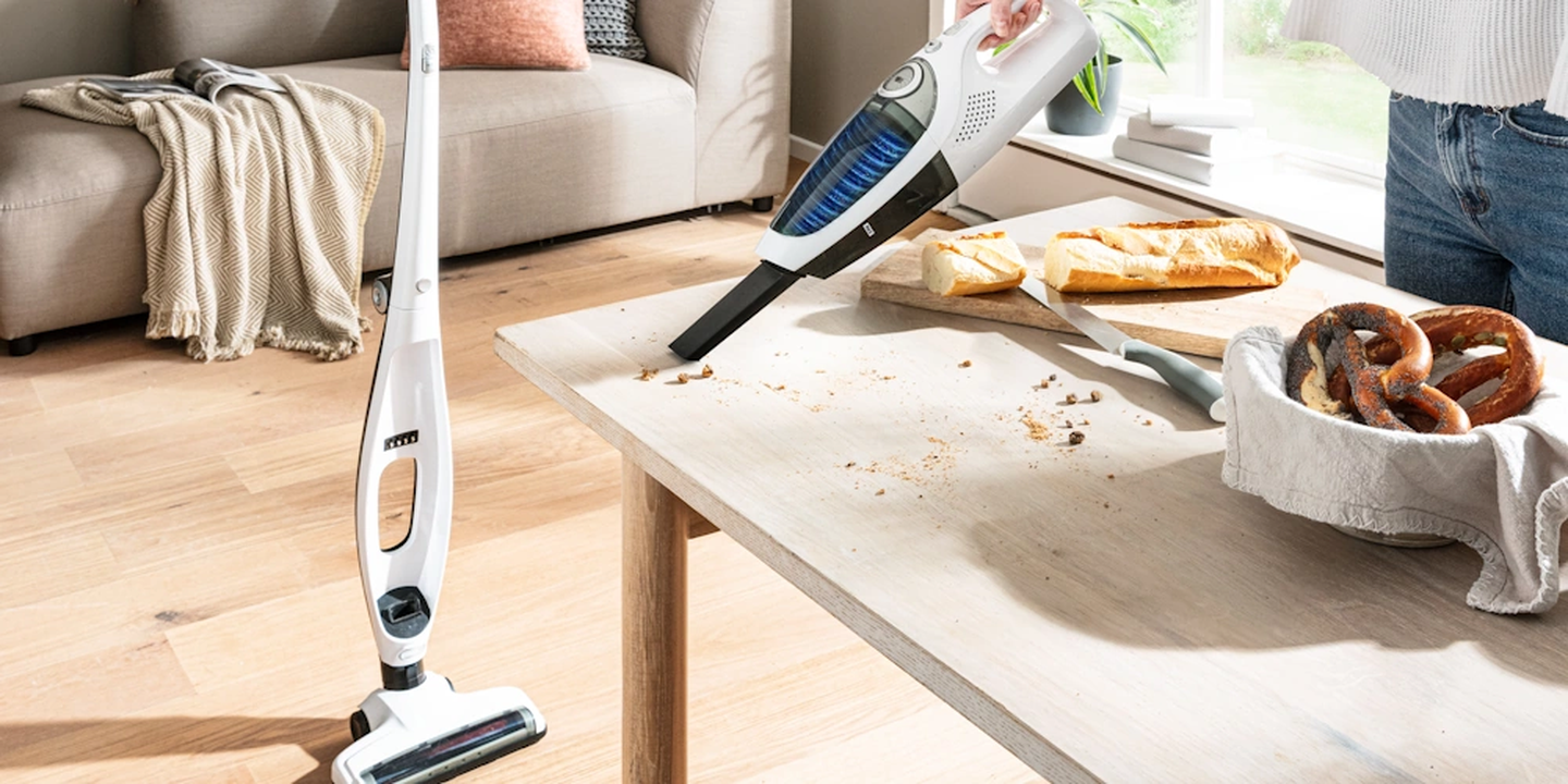 Eine Person saugt mit einem Handstaubsauger Krümel von einem Holztisch in der Nähe von Brezeln auf, im Hintergrund sind ein Sofa und ein großes Fenster zu sehen.