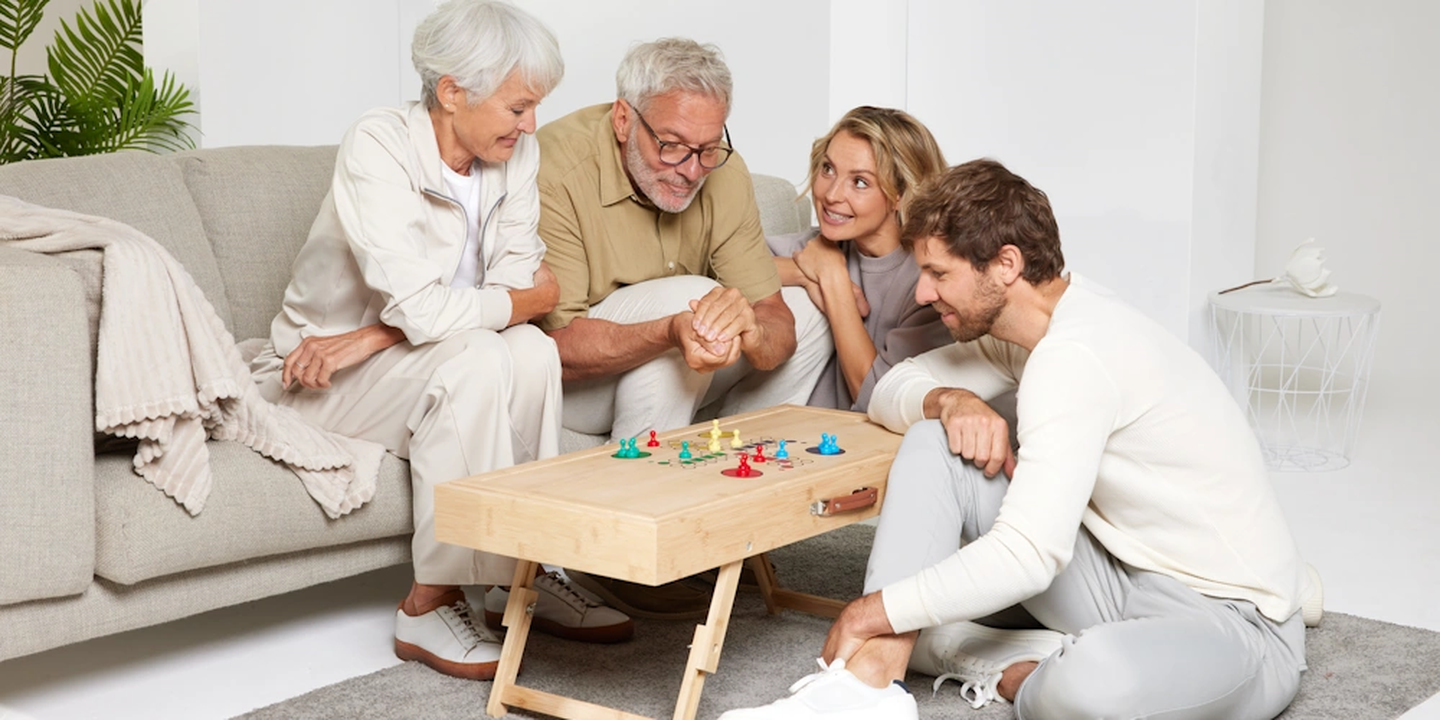 Zwei ältere Erwachsene und zwei jüngere Erwachsene sitzen um einen Puzzletisch herum, spielen ein Brettspiel und sitzen lächelnd und entspannt in einem gemütlichen Wohnzimmer.