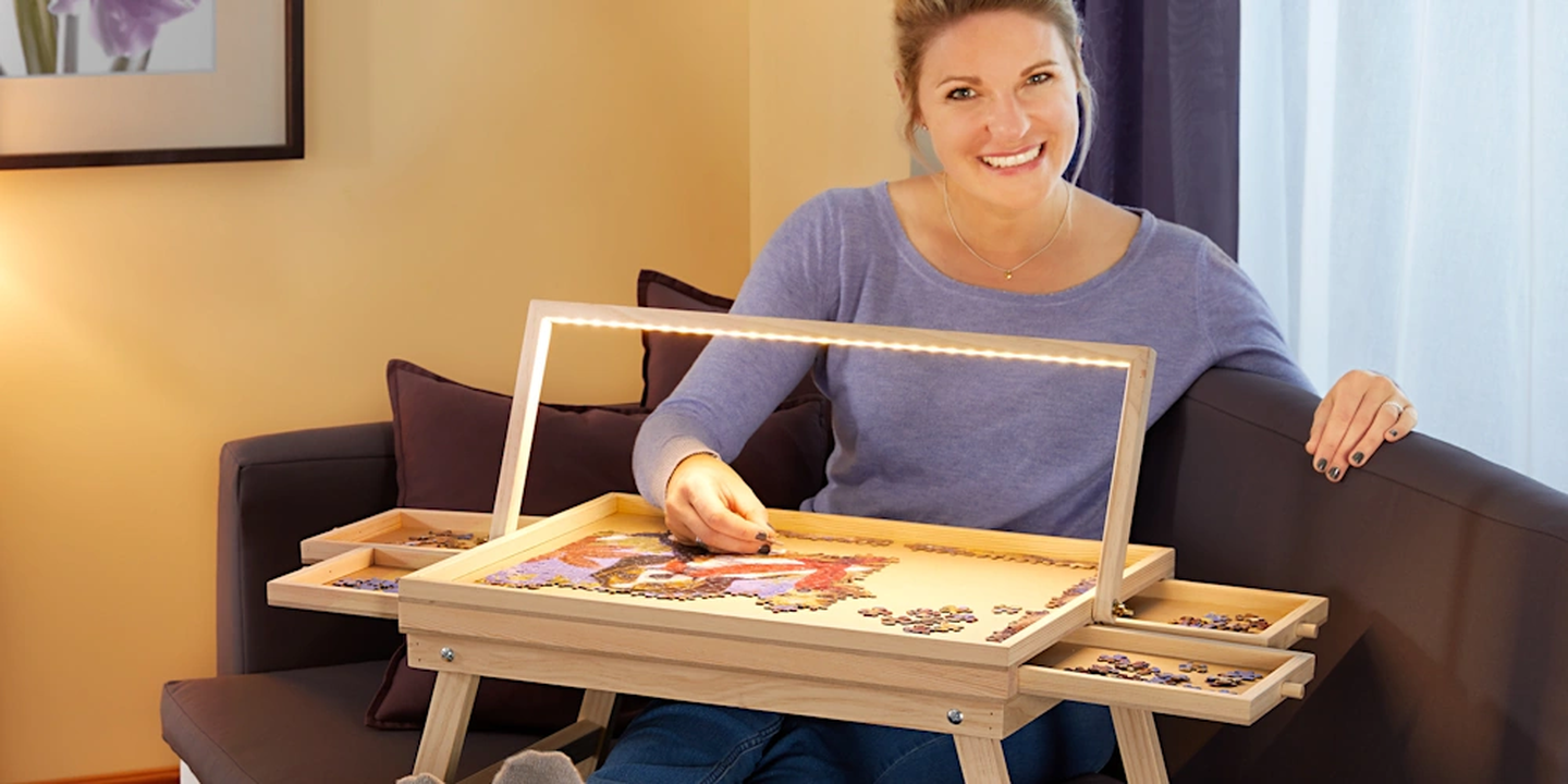 Eine lächelnde Frau, die auf einem Sofa sitzend ein Puzzle auf einem Puzzletisch mit LED-Beleuchtung zusammensetzt. Das Brett verfügt über Fächer für die Puzzleteile.