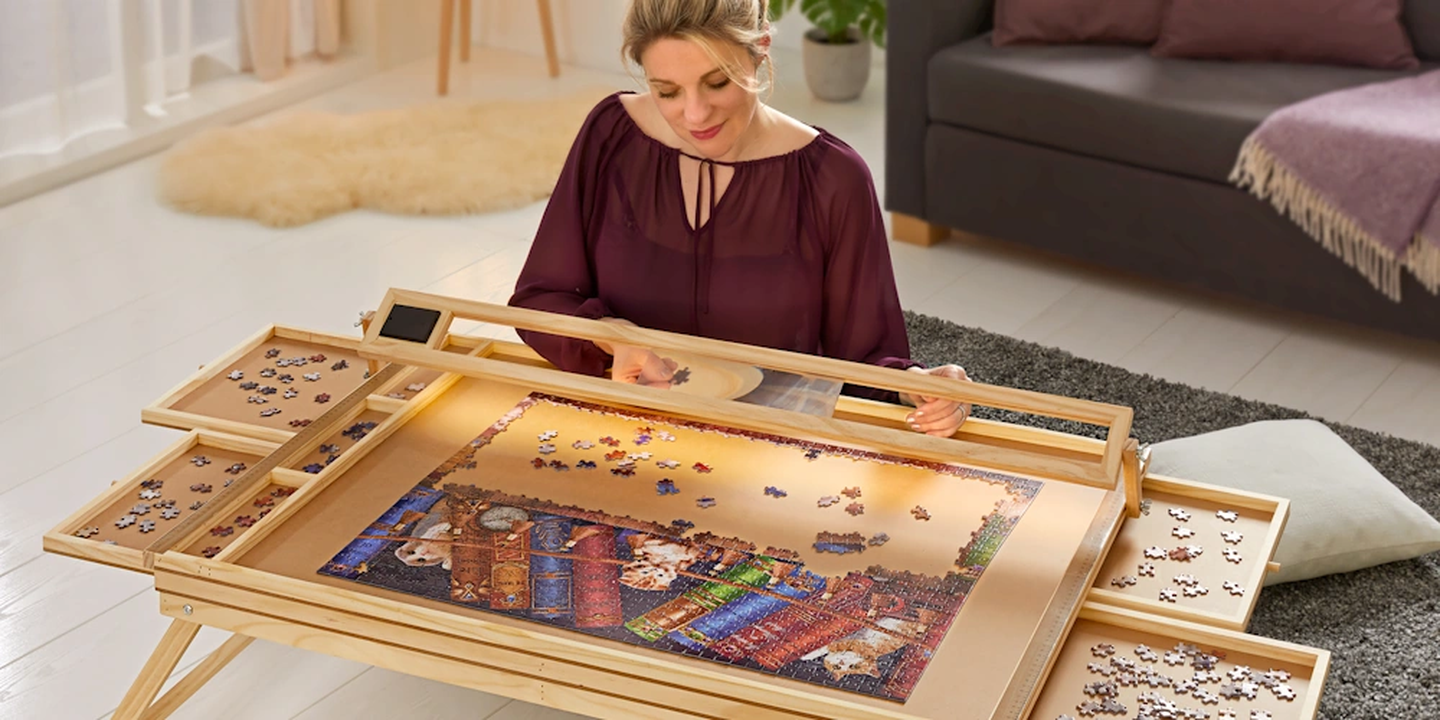Eine Frau, die ein buntes Puzzle auf einem hölzernen Puzzletisch mit ausziehbaren Schubladen zusammensetzt, während sie bequem in einem gemütlichen, gut beleuchteten Raum sitzt.