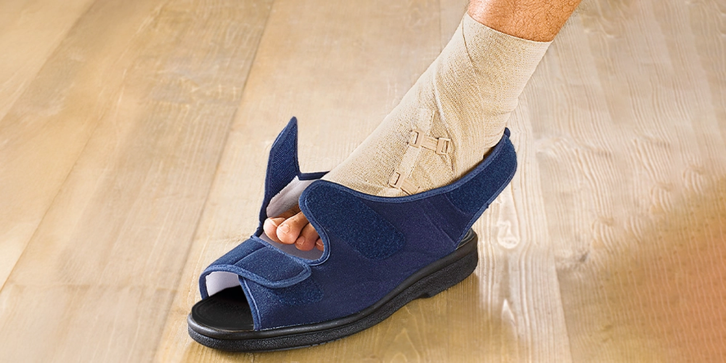 Ein Fuß, der mit einem beigen Verband umwickelt ist und einen blauen medizinischen Schuh mit verstellbaren Riemen trägt, steht auf einem Holzboden.
