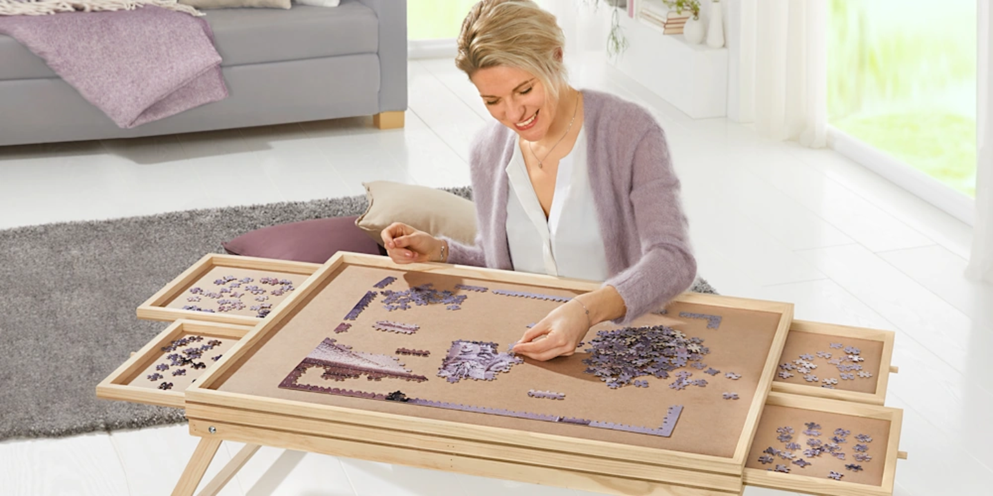 Eine Frau, die an einem hölzernen Puzzletisch mit ausziehbaren Ablagen ein Puzzle zusammensetzt, während sie in einem hellen, gemütlichen Wohnzimmer sitzt.